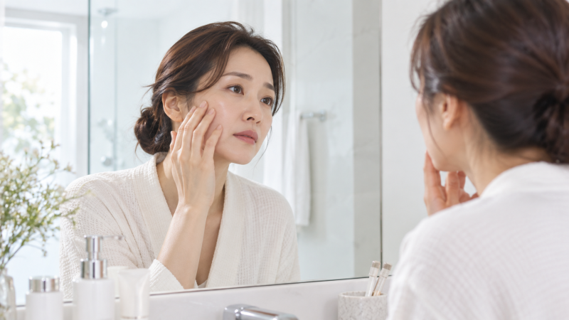 거울 앞에서 볼을 손으로 살피며 피부 상태를 확인하는 모습을 담은 이미지