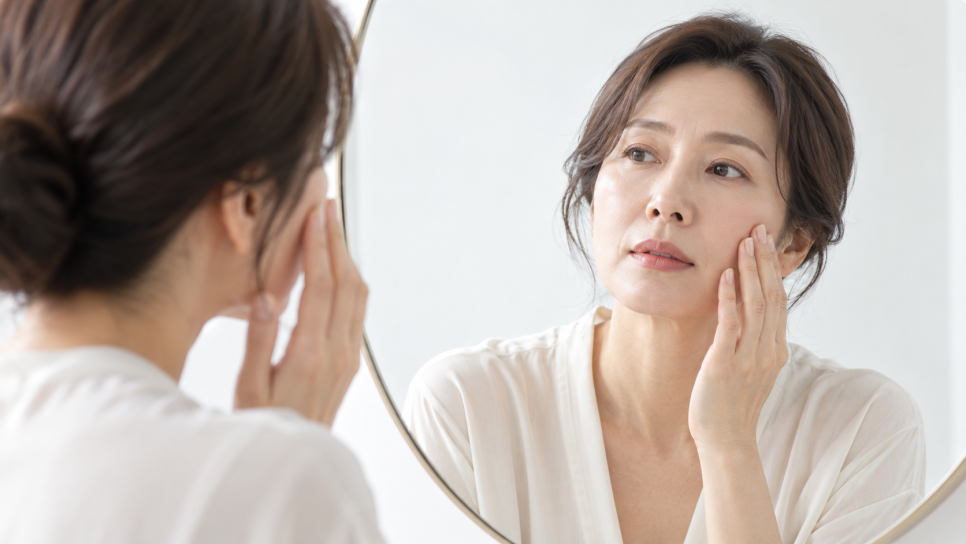 거울 앞에서 볼에 손을 대고 피부 상태를 살피는 여성의 모습을 담은 이미지